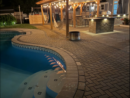 Nighttime backyard pool patio with pergola string lights, hanging hammocks, and outdoor kitchen and bar beside a house.