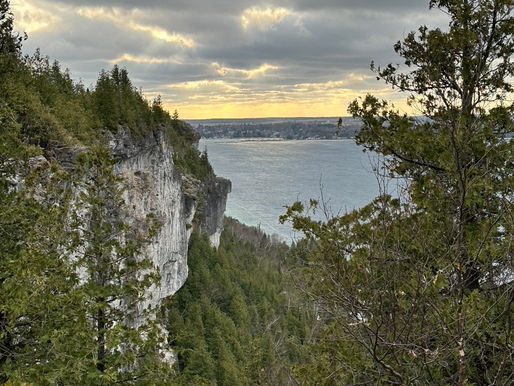 View of a dramatic cliff overlooking a calm lake, surrounded by dense green trees. Cloudy sky with a hint of golden sunset on the horizon.