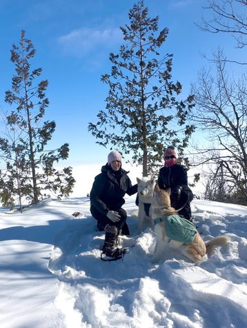 Two people in winter attire kneel in snow with two dogs, one wearing a green vest. Blue sky and trees in the background; a joyful scene.