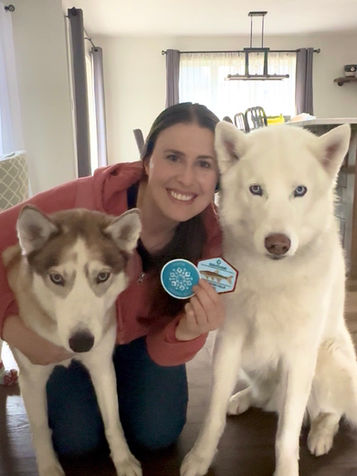 Smiling woman kneeling with two huskies indoors, holding blue badges. Bright room with a dining table in the background.