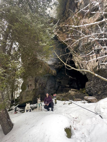 Woman with two dogs in snow at cave entrance. Pine trees and rock formations in the background. Warning sign nearby. Winter scene.