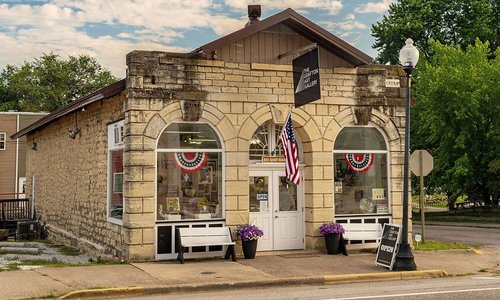 Stone-front art gallery on Main St with U.S. flag, open sign, benches and flowers under a cloudy sky.