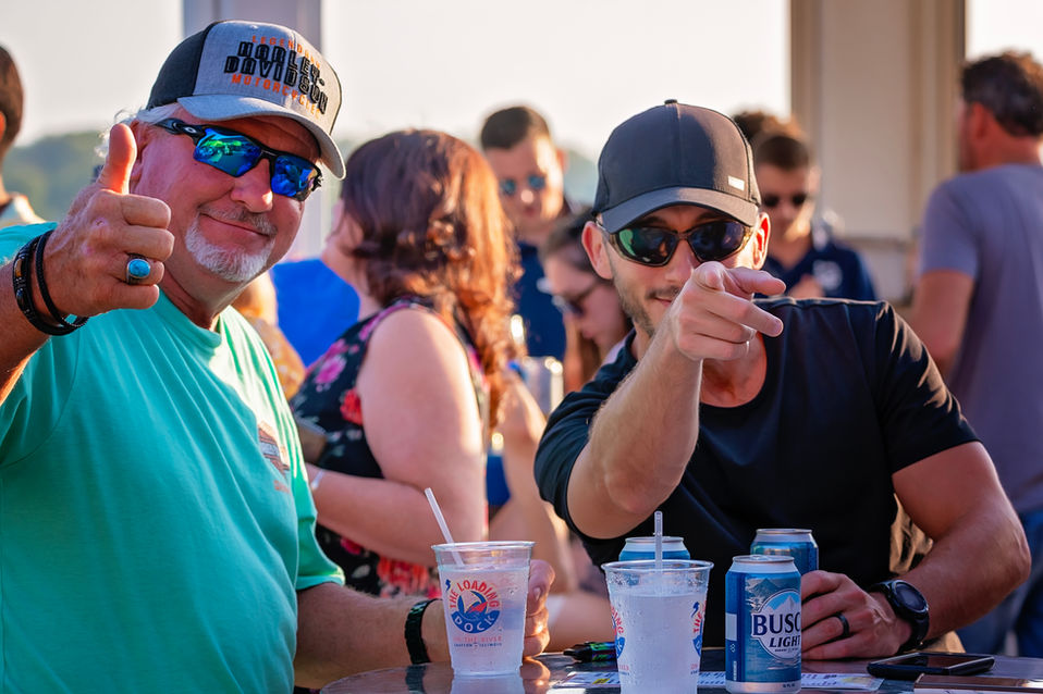 Two men with drinks and sunglasses are pointing and smiling.