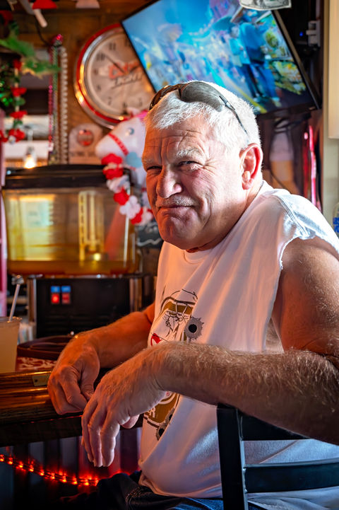 Smiling older man at a bar, wearing sunglasses, enjoying a drink.
