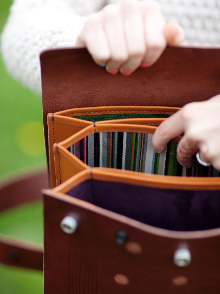 The inside of a brown leather bag, which has concertinaed pockets inside, and is lined with striped material.