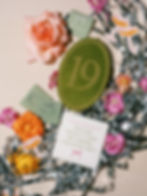 Wedding Invitation flatlay photographed by Ruet Photo. Green ribbon and individual pink, purple, and orange florals surround the green velvet table number, drink coaster reading "Please Don't Take My Drink, I'm Dancing!", die-cut custom coupe glasses, and matchbooks.