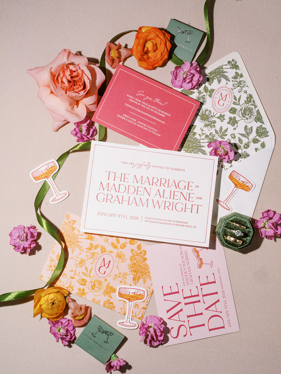 Wedding Invitation flatlay photographed by Ruet Photo. Green ribbon and individual pink, purple, and orange florals surround the wedding invitation, details card, save the date, die-cut custom coupe glasses, and matchbooks. 