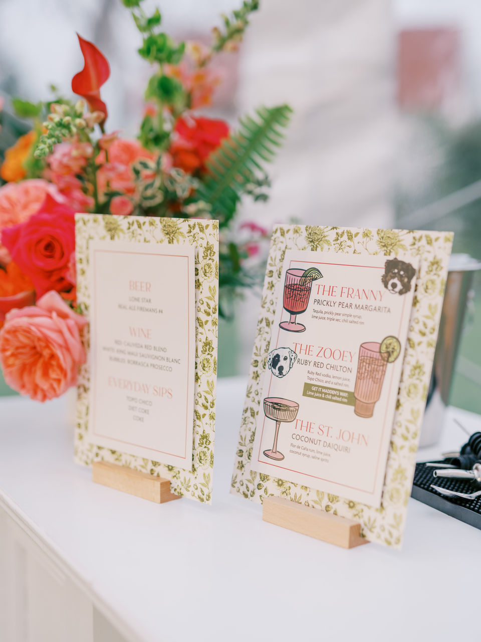 Wedding Signature Drinks Menu and Bar Menu on a white bar next to a colorful pink and orange floral arrangement. Green and white floral backgrounds on the menus, with custom illustrations for drinks, "The Franny - Prickly Pear Margarita," "The Zooey - Ruby Red Chilton," and "The St. John - Coconut Daiquiri."