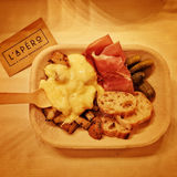 A to go container on a table with melted raclette cheese over potatoes, cured meats, cornichons and baguette.