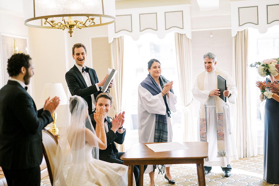 Bride and groom seated, clapping alongside standing guests wearing formal attire in a bright room. Joyful wedding ceremony atmosphere.