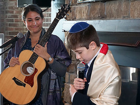 An officiant with a guitar and a boy in a kippah and shawl hold microphones, standing by a brick wall. The mood is solemn and respectful.
