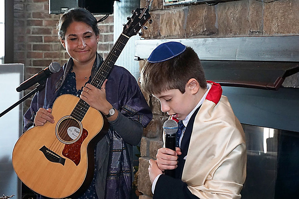 An officiant with a guitar and a boy in a kippah and shawl hold microphones, standing by a brick wall. The mood is solemn and respectful.