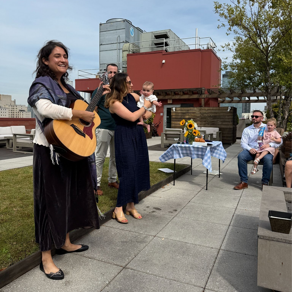 Smiling cantor plays guitar at rooftop baby naming ceremony. Baby held by another woman. Table with sunflowers and cake nearby. Relaxed, sunny setting.
