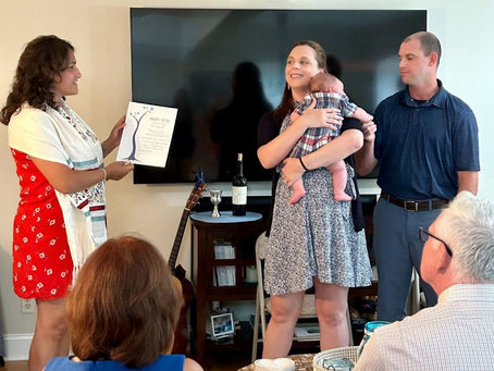 A Jewish officiant in red dress holds a certificate, smiling at a couple with a baby. A guitar and wine on a table in home setting. Mood is celebratory.