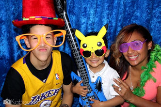 Three people in photo booth with props: top hat, Pikachu, guitar