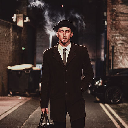 night portrait in nottingham, united kingdom of a man in a mafia style old school suit smoking a cigar with smoke in the background in an alleyway with a blacked out range rover in the background