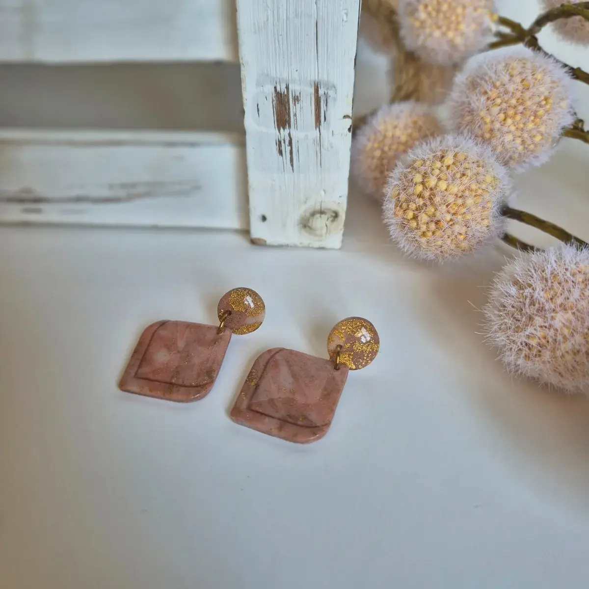 Blush pink marbled clay earrings with layered square drop design and shimmering gold leaf accents, styled on neutral fabric