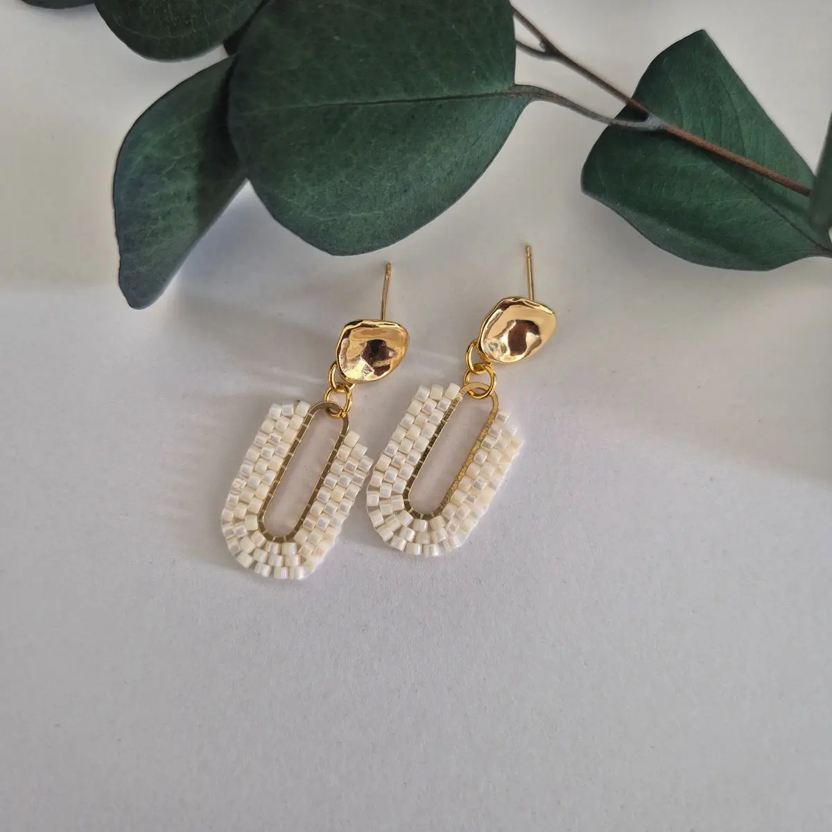 Glistening Sands ivory beaded drop earrings with gold hammered studs displayed on a white background with eucalyptus leaves.