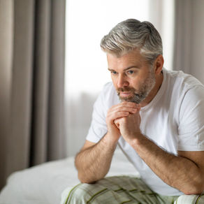 Thoughtful middle aged gay man in a white shirt sits on a bed, hands clasped, wondering about how to protect his brain health and reduce risk of dementia.