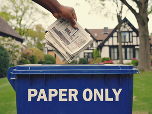 A clean and eco-friendly suburban New Jersey neighborhood on a sunny day, with blue recycling bins on the curb, labeled 'Paper Only.' A hand is placing newspapers and flattened cardboard into the bin.