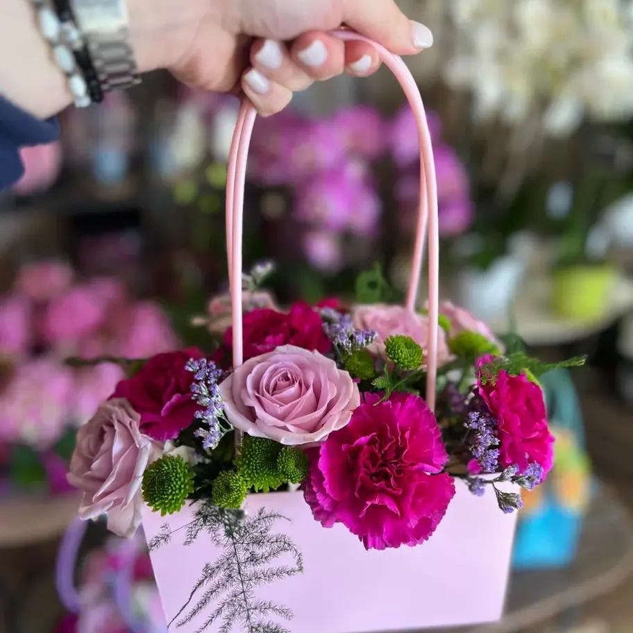 Une main tient un petit panier de fleurs roses et fuchsia, comprenant des roses, des œillets et des petites fleurs vertes.