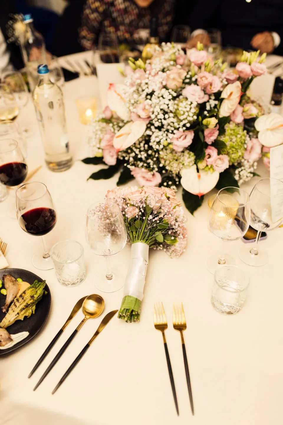 Table dressée pour un repas festif avec un centre de table floral composé de roses roses, d'anthuriums et de gypsophiles, des verres de vin rouge, des couverts dorés et une assiette de légumes.