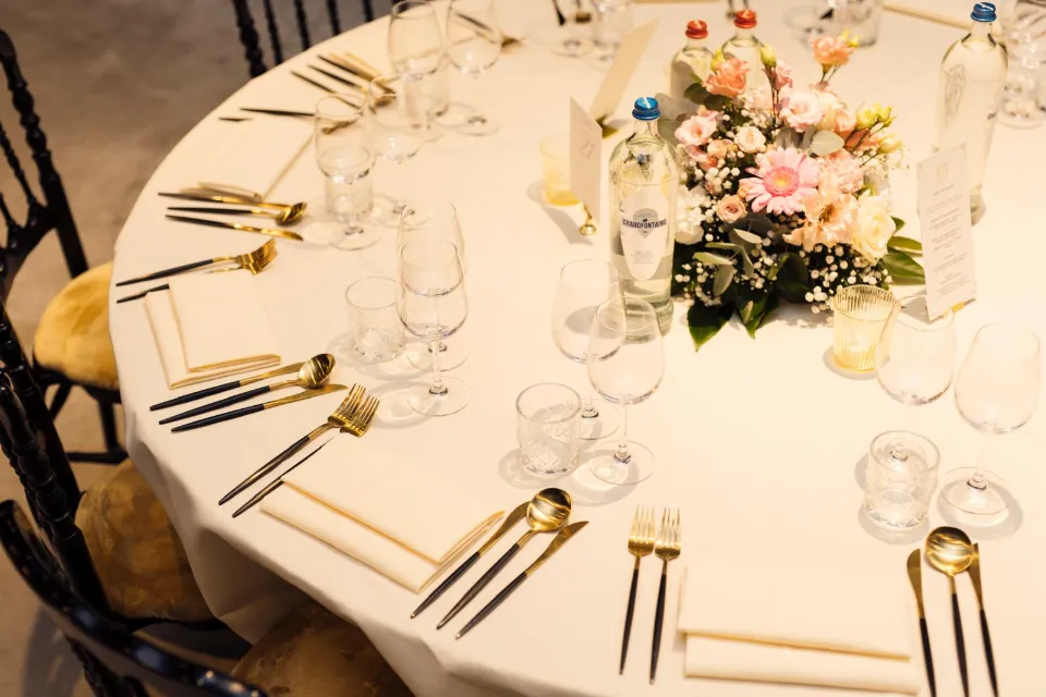 Table de mariage dressée avec des serviettes en tissu, des couverts dorés et noirs, des verres à vin transparents et un bouquet de fleurs roses et blanches.