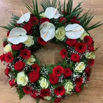 Couronne funéraire circulaire composée d'anthuriums blancs, roses rouges, gerberas rouges et feuillage vert.