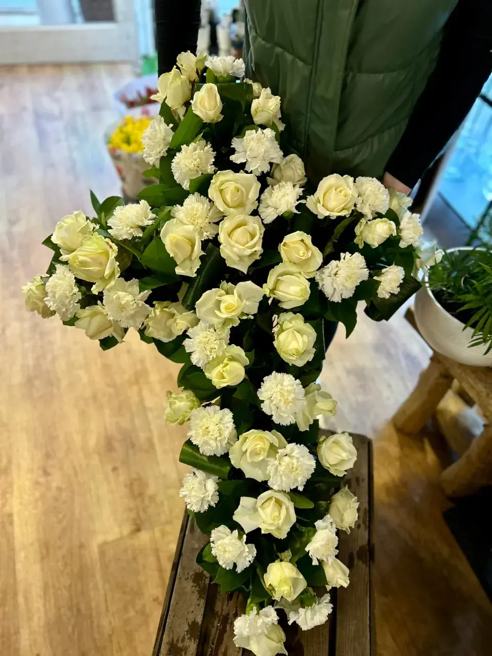 Un arrangement floral en forme de croix, composé de roses blanches et de petits œillets blancs, sur un fond en bois.