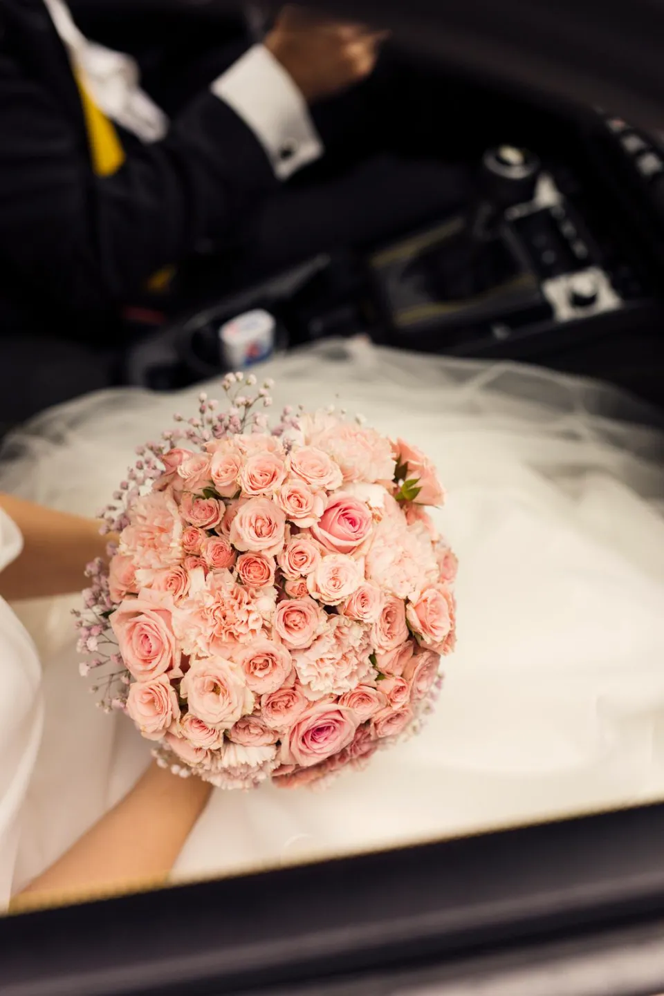 Un bouquet de roses roses et de gypsophiles porté par une mariée, avec un homme en costume en arrière-plan