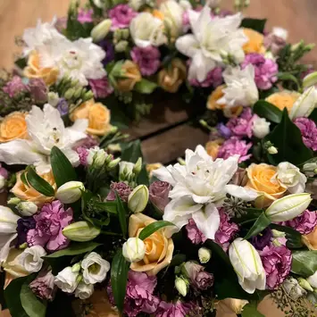 Couronne de fleurs composée de roses saumon, de lys blancs et de fleurs roses sur fond boisé.