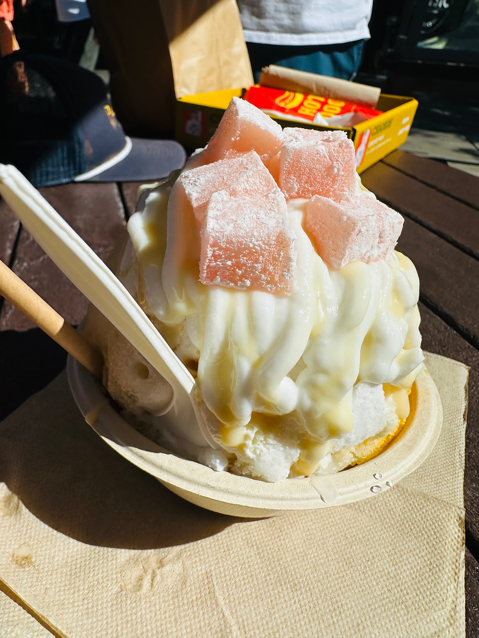 Shaved ice dessert topped with peach cubes and creamy sauce in a paper bowl, served outdoors on a table with a spoon and snack box