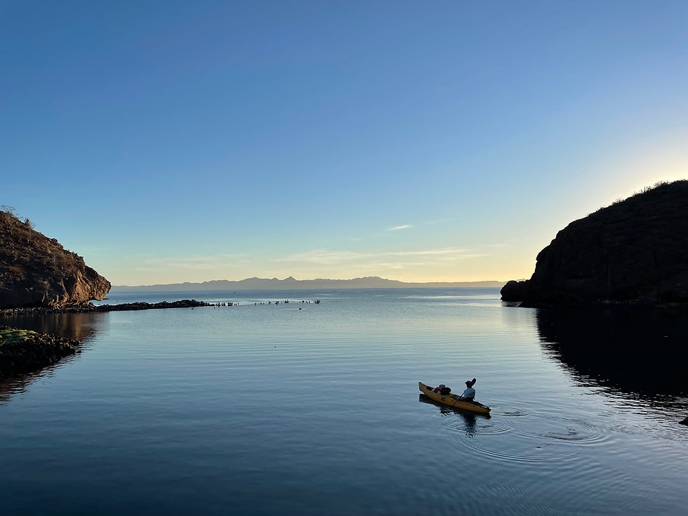 Lone kayaker in a yellow kayak on calm water between dark cliffs at sunrise, with distant mountains and a clear blue sky.