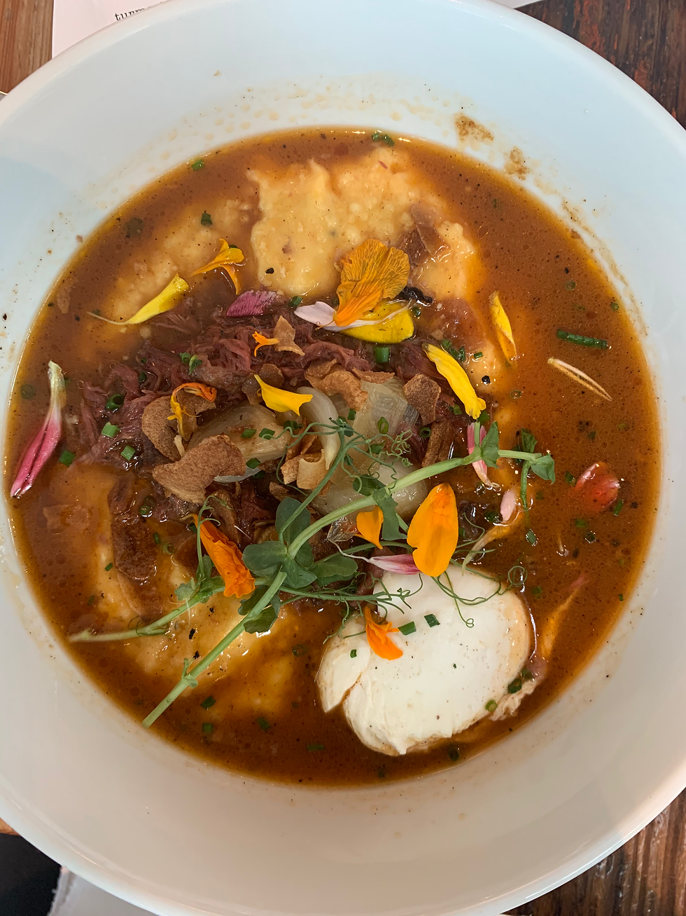 A white bowl of rich, savory stew served over grits, topped with shredded braised meat, a poached egg, green microgreens, and bright edible flower petals.