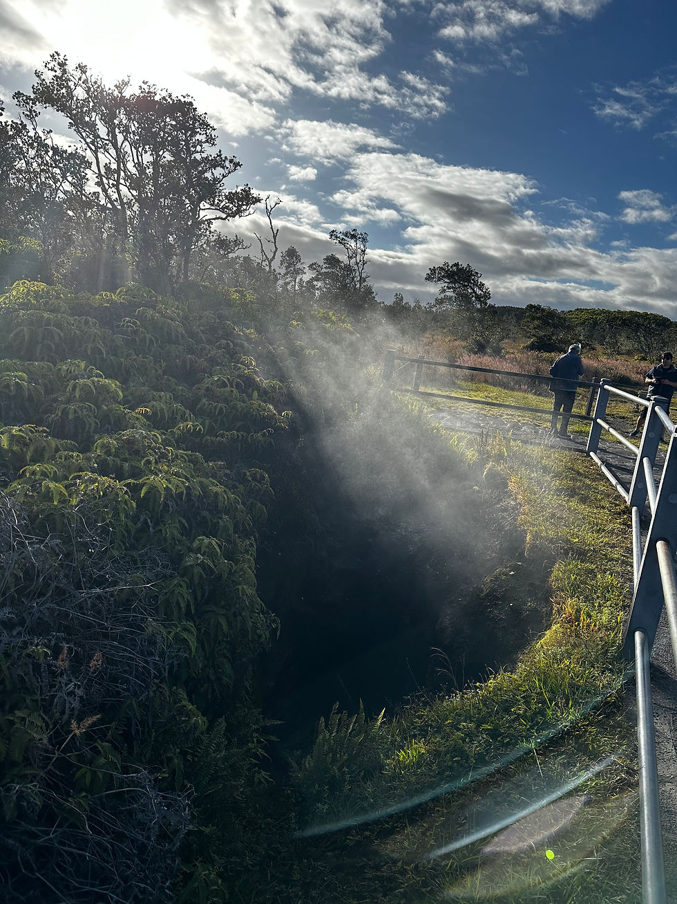 Sunlit mist rises from a dark volcanic crater at a lookout, with two people by a metal railing under a blue, cloudy sky.