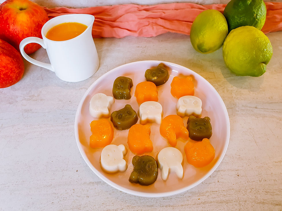 A pink plate with colourful, animal shaped real fruit jellies on it. Next to the plate is a jug of orange juice and some apples and limes.