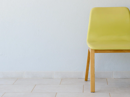 Une chaise moderne au dossier et à l'assise en plastique moulé de couleur jaune citron repose sur un sol carrelé clair, adossée à un mur blanc texturé. Ce siège vide et isolé symbolise l'absence criante des femmes handicapées dans les stratégies d'inclusion, laissant une place qui devrait être occupée mais qui reste inexploitée.