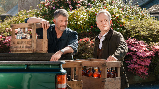 "Rosamunde Pilcher: Wo Liebe aufhört und wo sie beginnt": Vor einer blühenden Hecke: Andrew Evans (Simon Böer) und Rodney Cumberwell (Ian Dickinson) stehen an der Ladefläche eines grünen Pickups. Beide haben rustikale Holzkisten mit gefüllten Glasflaschen vor sich stehen. Sie blicken aufmerksam auf etwas an der Seite.