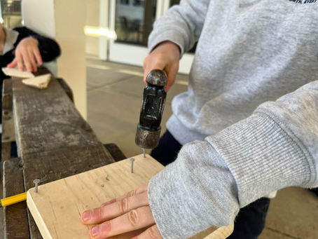 Close-up of a child's hands using a hammer and nail on a wooden plank during an Electivity woodworking enrichment class