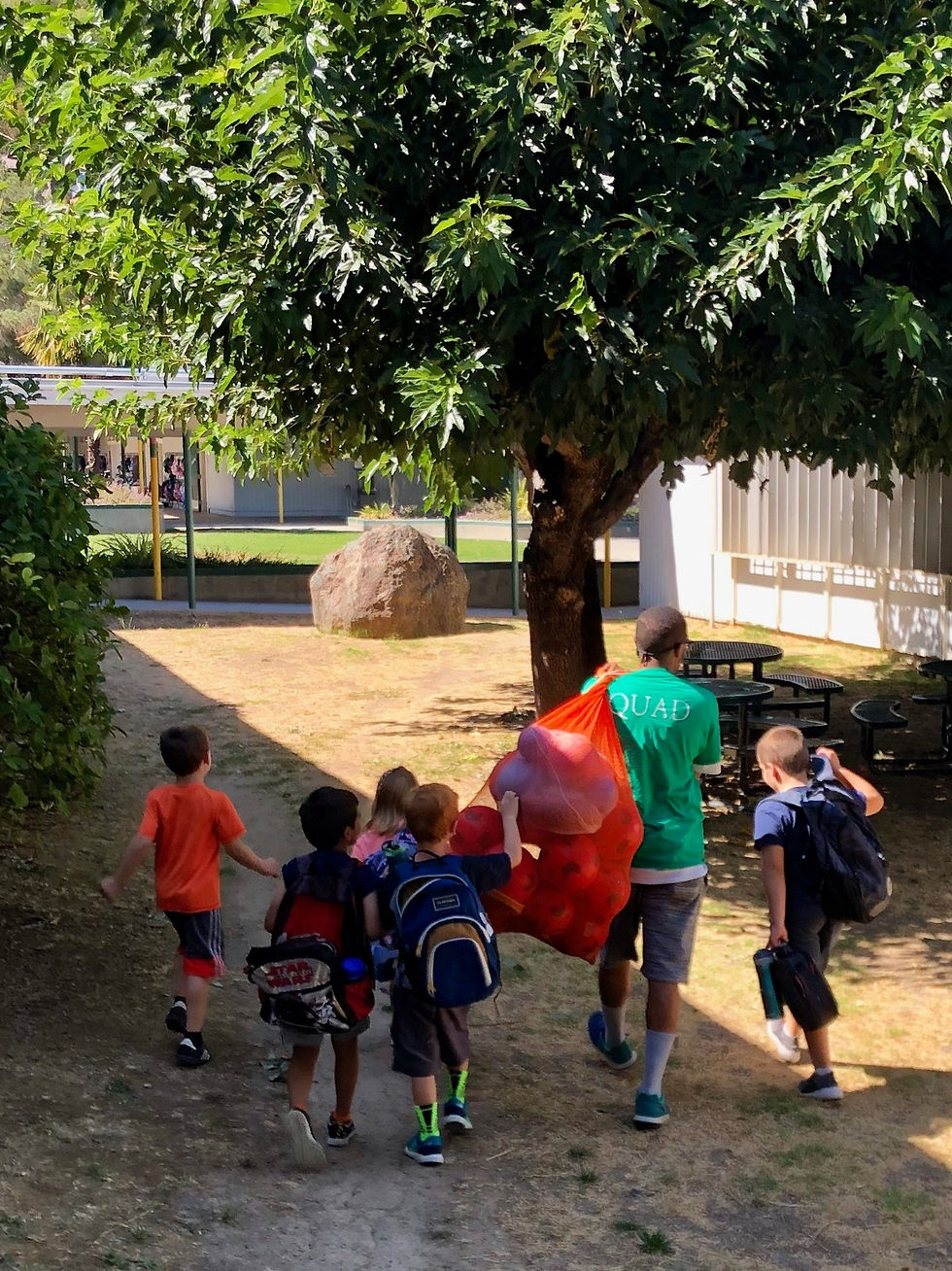 A group of young students seen from behind, walking across toward a sports field. They are following a soccer instructor who is carrying a large mesh bag filled with soccer balls over their shoulder.
