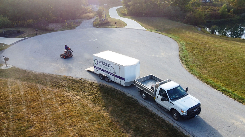 Truck towing a Werle's Lawn Service trailer on a paved road, man riding a lawnmower nearby. Sunny day, grassy surroundings.