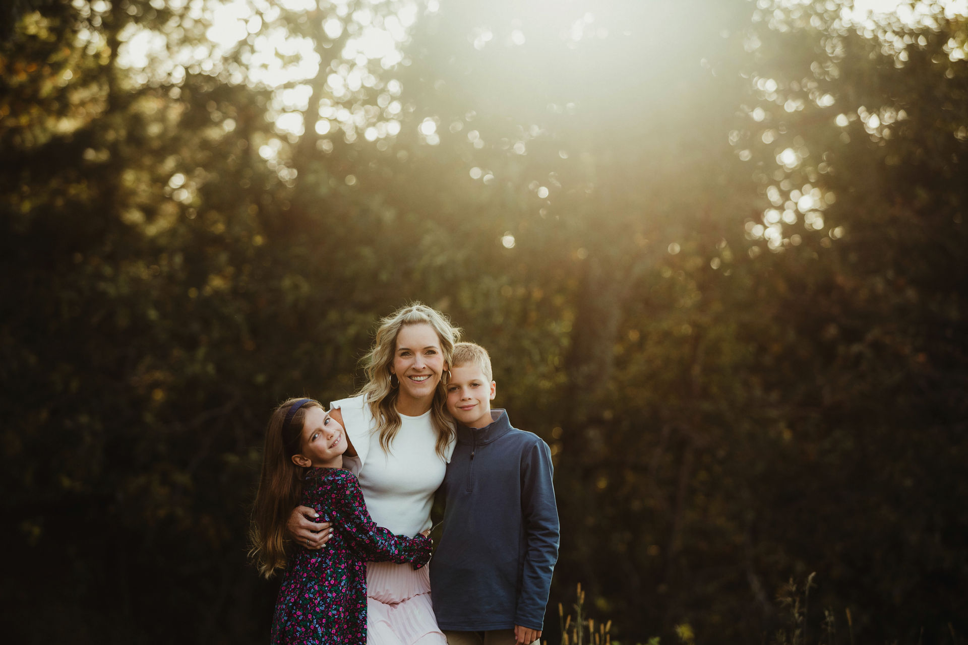 Mom standing in the middle of her son and daughter hugging with the warm sun behind them.