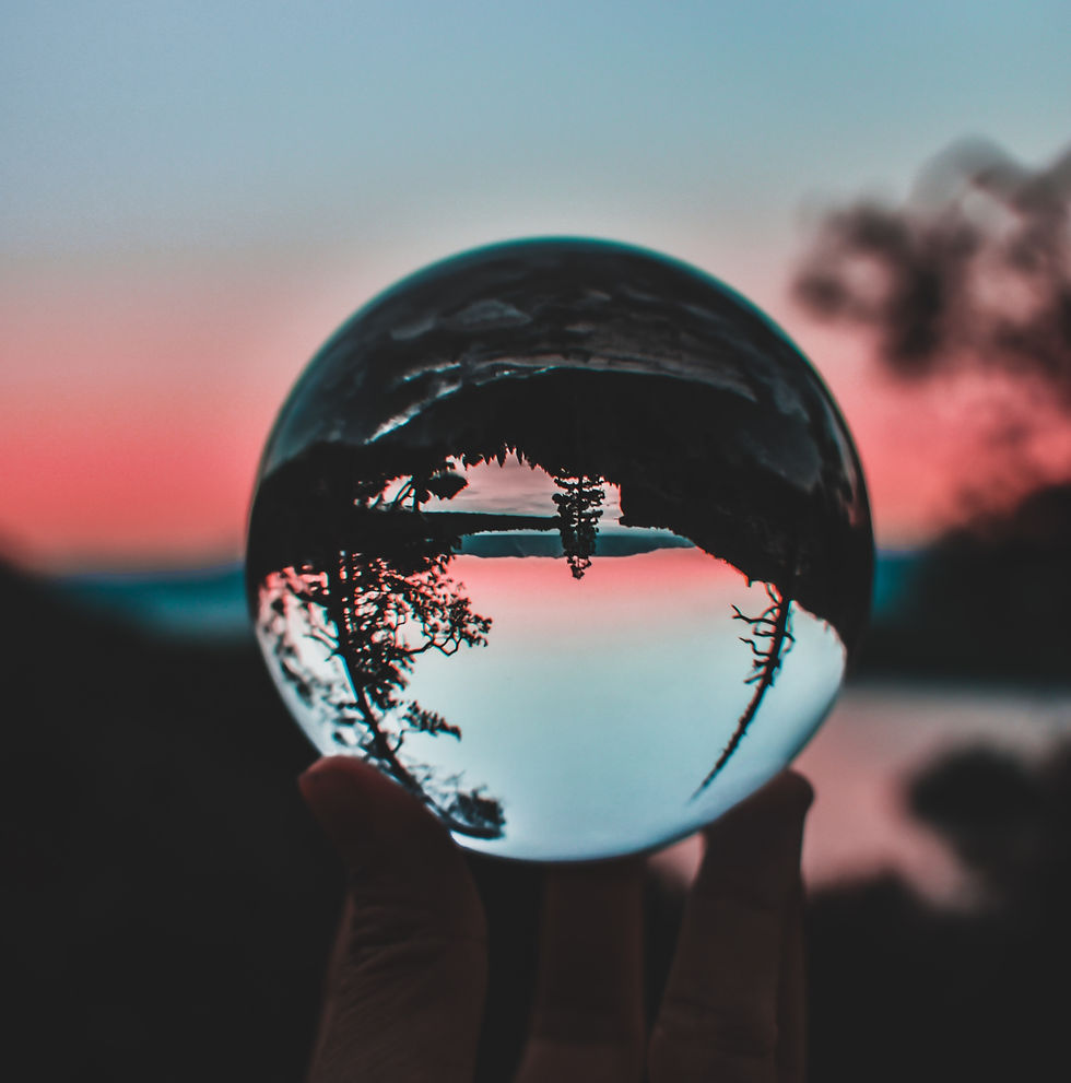 An image of a clear crystal sphere being help up showing a inverted reflection in the sphere of a forest landscape. There is a sunset in the background.
