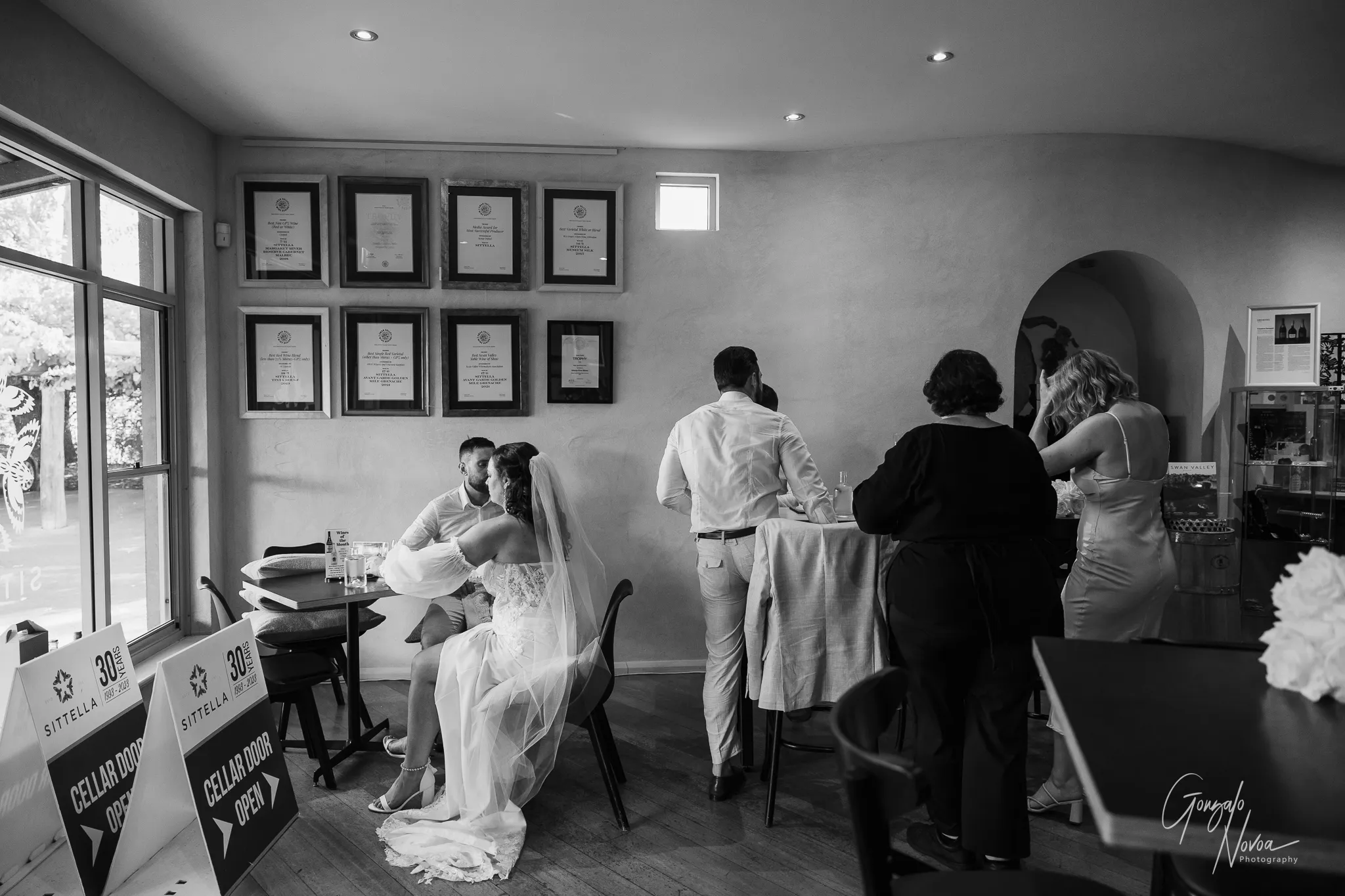 A black and white image of newlyweds Tahnee and James sharing a private moment seated inside the Sittella Winery cellar door, surrounded by friends and framed wine awards on the wall