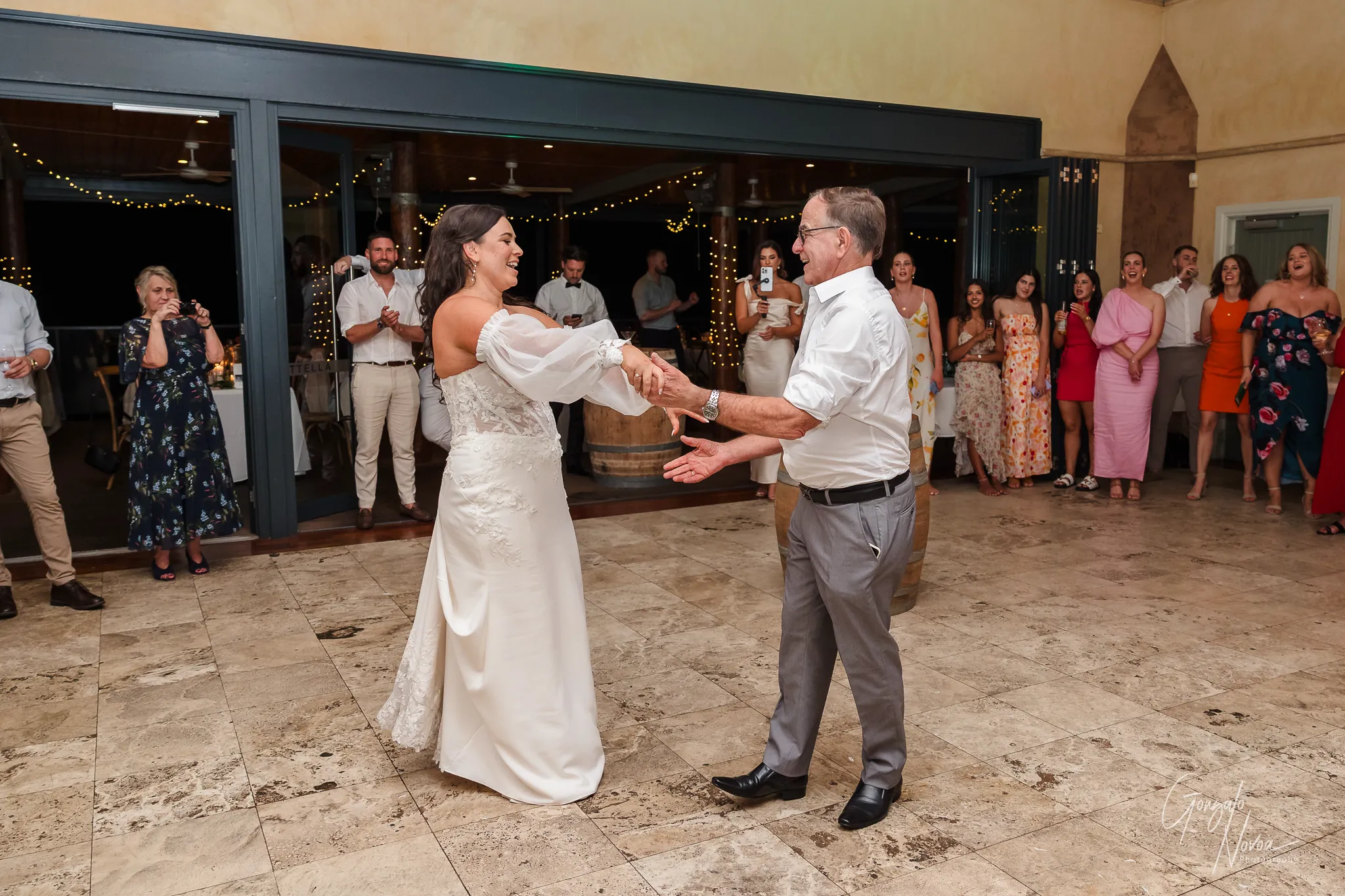 The bride and her father dance together during the wedding reception, laughing and holding hands in a lively, happy moment. The bride wears a strapless wedding gown with sheer off-shoulder sleeves, and her father is dressed in a white shirt and grey trousers. In the background, guests cheer them on, some taking photos and videos, while fairy lights add a festive touch to the evening atmosphere.