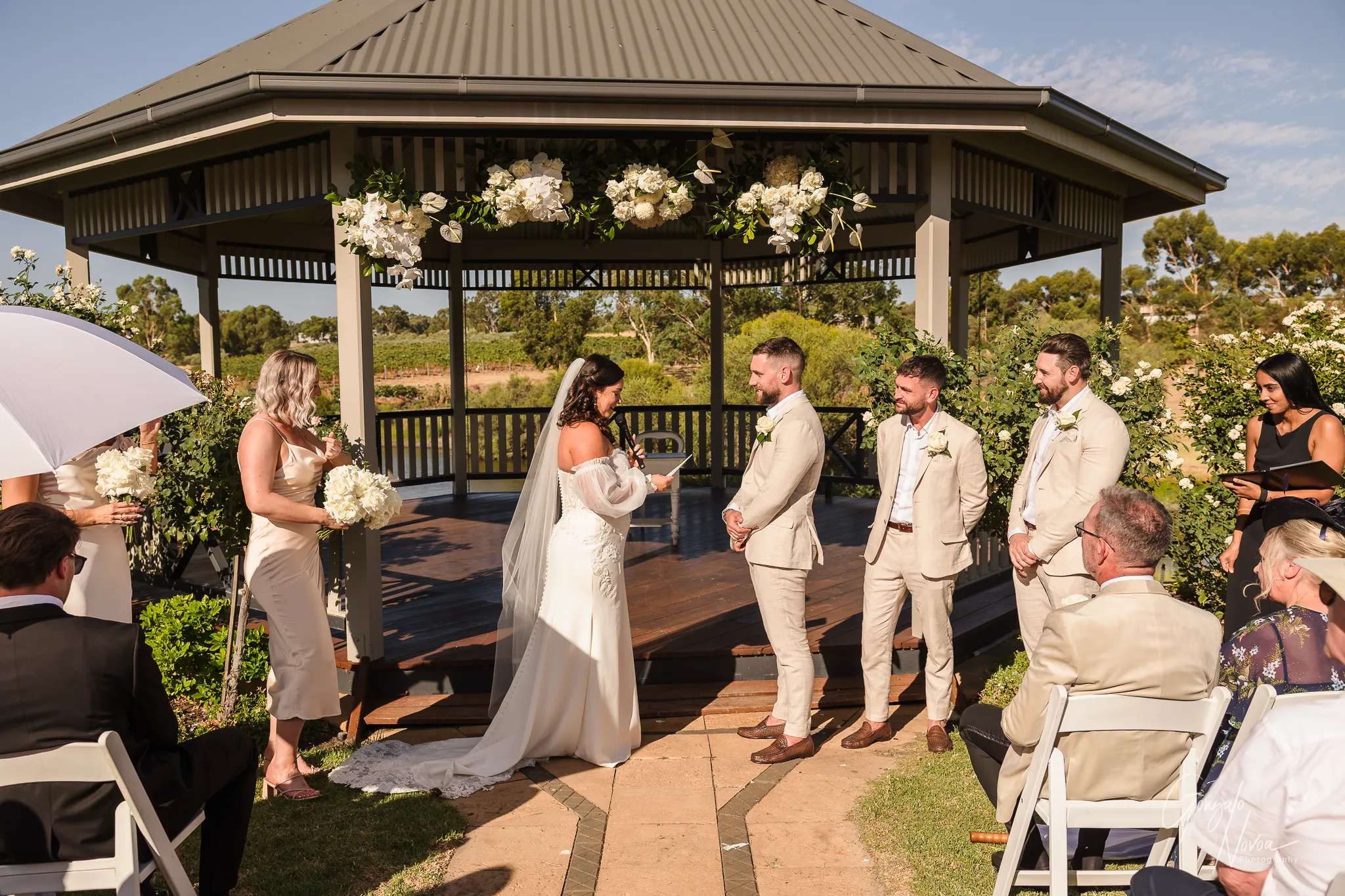 Bride Tahnee standing under a floral-decorated gazebo at Sittella Winery, reading her vows to groom James as their bridal party and guests watch on during a sunny outdoor ceremony.