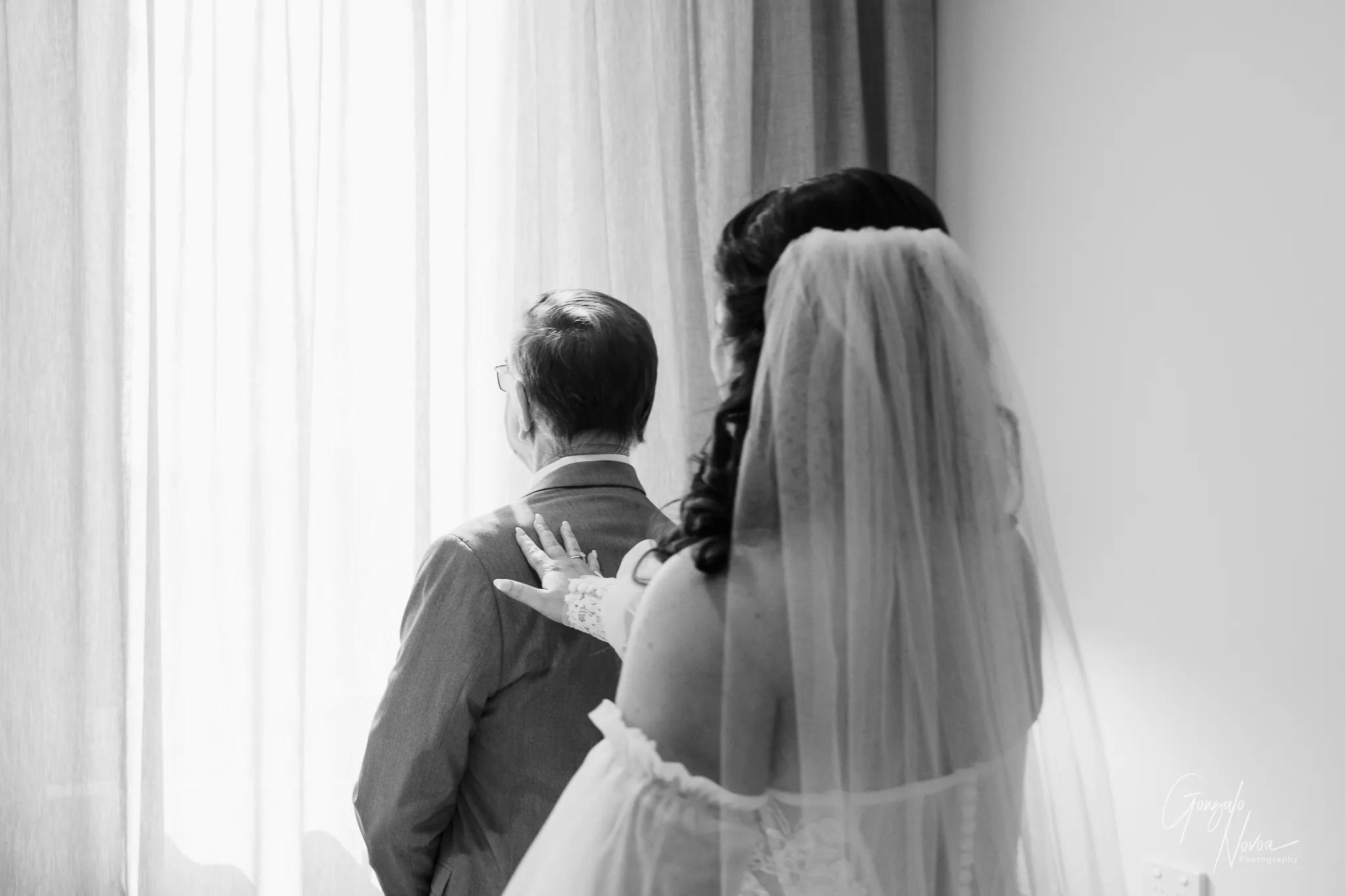 Black and white photo of bride Tahnee reaching out to tap her father’s shoulder during their emotional first look, as he stands facing a window with sheer curtains letting in soft light.