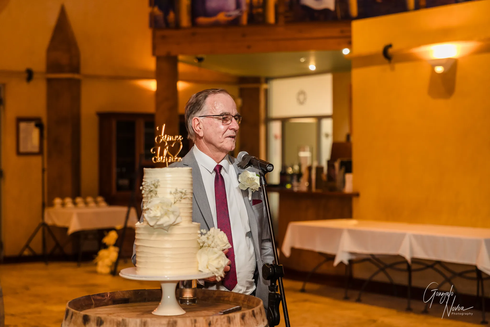 The bride's father wearing a grey suit and red tie gives a heartfelt speech at the wedding reception, standing beside a white tiered cake adorned with flowers. The scene is warmly lit, capturing the emotional and celebratory atmosphere inside the venue.