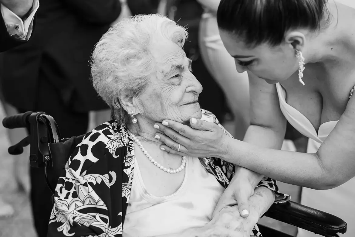 A heartwarming black-and-white photo of bride Maryanne gently touching her grandmother’s chin during her wedding at Our Lady of the Rosary Parish, Woodlands, WA. The bride’s grandmother is seated in a wheelchair, smiling softly as they share this intimate, emotional moment. The bride wears elegant earrings and a white dress, while her grandmother is adorned in pearls and a patterned jacket.