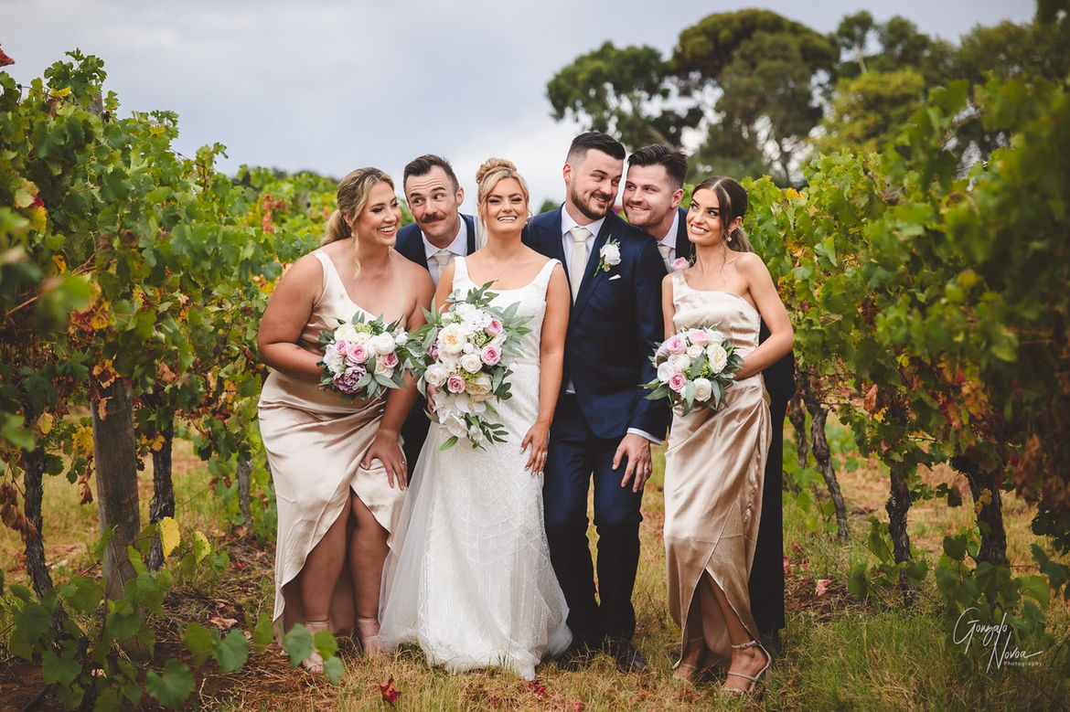 Bella and Liam pose with their bridal party in the vineyards at Sandalford Wines, Caversham WA. The bride is in a white gown, and the bridesmaids wear champagne-coloured dresses, while the groom and groomsmen are in navy suits. The group is smiling and leaning in close, creating a joyful and lighthearted scene among the vines.
